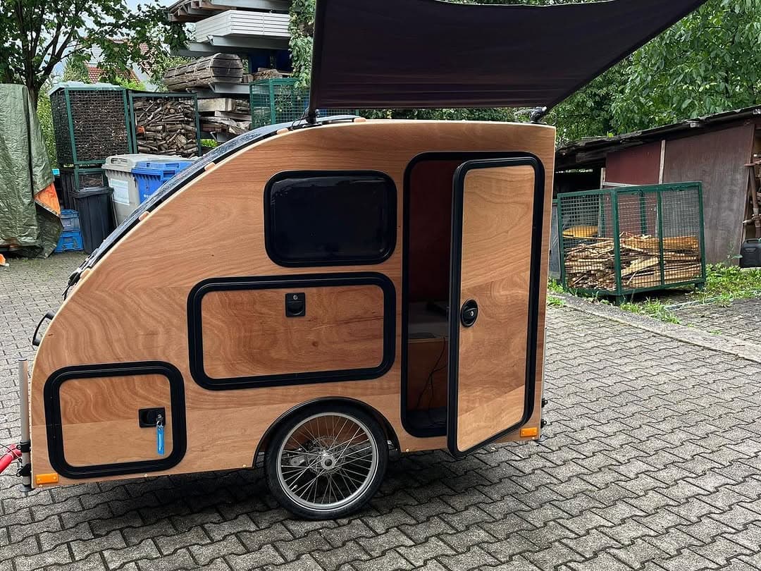 Fahrrad-Camper i:woody aus Holz mit geöffneter Tür und aufgespanntem Sonnensegel